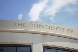 Photograph of the university of sign on top of arched office building with sky above. Windows visible below sign. Image taken on the campus of a public four year university in the United States.